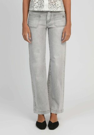 Femme portant un jean gris clair à jambes droites avec des perles en décoration sur le côté et des ballerines noires pointues, se tenant devant un fond uni.