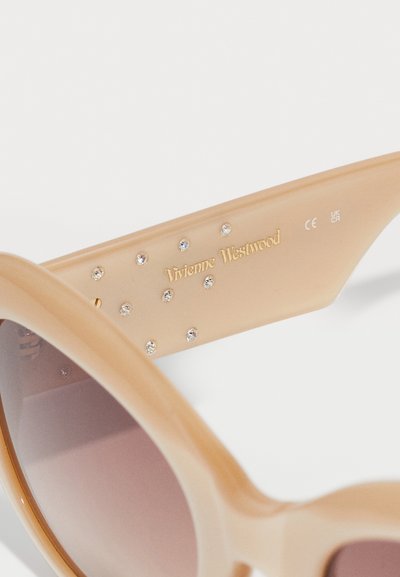 Beige γυαλιά ηλίου με γυαλιστερή επιφάνεια, διαθέτοντας gradient φακούς και κρυστάλλινες λεπτομέρειες ενσωματωμένες στο βραχίονα. Χρυσό κείμενο logo εμφανίζεται.