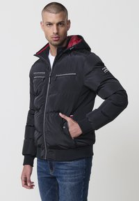 Svart puffjacka med röd foder, utrustad med dragkedjeförsedda bröstfickor, ribbade ärmmuddar och en huva med dragsko. Stylad med blåjeans.