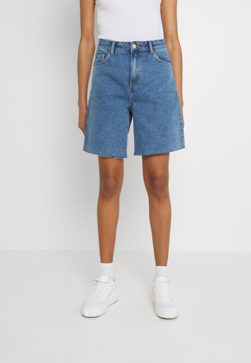 Personne portant un short en jean bleu mi-long, un débardeur blanc, des chaussettes blanches et des baskets blanches, debout devant un fond blanc uni.