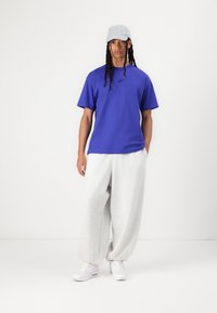 T-shirt royal blue à manches courtes avec un logo noir, associé à un pantalon de jogging baggy gris clair et des baskets blanches. Accessoirisé d'une casquette gris clair.