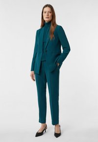 Turkusowy garnitur uszyty z dopasowanej tkaniny, zawierający dopasowaną marynarkę i zwężane spodnie. Połączony z czarnymi szpilkami z ostrym noskiem.
