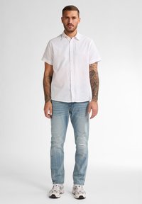 Korte mouwen, wit linnen overhemd met kraag, gecombineerd met lichtblauwe versleten jeans en witte sneakers met grijze accenten.