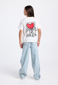 Wit katoenen T-shirt met een grafisch ontwerp van twee figuren die een rood hart vasthouden, gecombineerd met lichtblauwe wijde jeans.