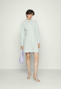 Vestido camisola em xadrez verde e branco com mangas longas, fecho de botão, cintura ajustada e saia rodada; combinado com uma mala lilás e sandálias.