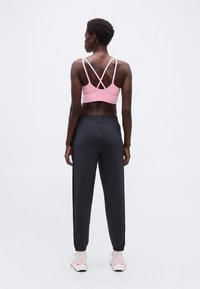 Γυναίκα που στέκεται με την πλάτη στην κάμερα, φορώντας ροζ σπορ σουτιέν με διασταυρούμενους ιμάντες, μαύρο παντελόνι jogger, λευκές κάλτσες και λευκά αθλητικά παπούτσια με ροζ λεπτομέρειες.