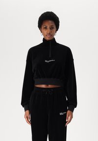 Black corduroy zip-up sweatshirt with a cropped design; features long sleeves, a high collar, and embroidered logo. Paired with matching pants.