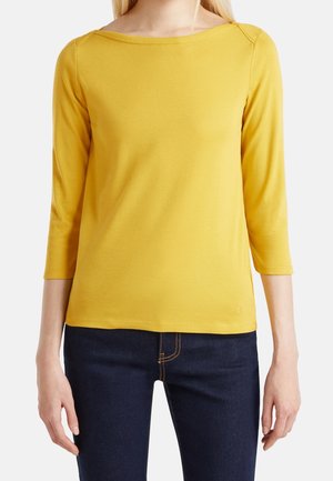 Femme portant un haut jaune uni à manches longues avec encolure bateau et un jean bleu foncé visible des épaules aux hanches.