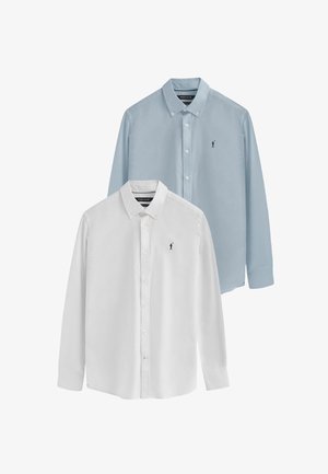 Biała koszula z długim rękawem, zapinana na guziki, z małym logo na piersi, w połączeniu z ciemnoszarymi spodniami i jasnobeżowymi sneakersami. Prosty krój i dopasowanie.