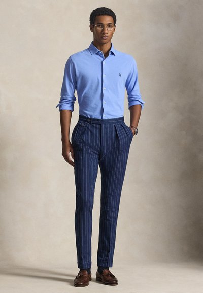 Άνδρας με γυαλιά, ανοιχτό μπλε πουκάμισο με κουμπιά, σκούρο μπλε παντελόνι με ρίγες, καφέ μοκασίνια, με τα χέρια στις τσέπες, στέκεται μπροστά σε απλό φόντο.