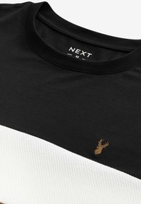 Schwarzes T-Shirt mit kurzen Ärmeln und weißen strukturierten Streifen, mit einem goldenen gestickten Hirschlogo nahe dem Saum. Glattes Material, Rundhalsausschnitt.