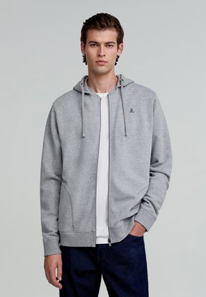 Felpa grigia con zip e cappuccio regolabile, polsini a costine e tasche. Indossata sopra una T-shirt bianca e abbinata a jeans scuri. Dettaglio del logo semplice.