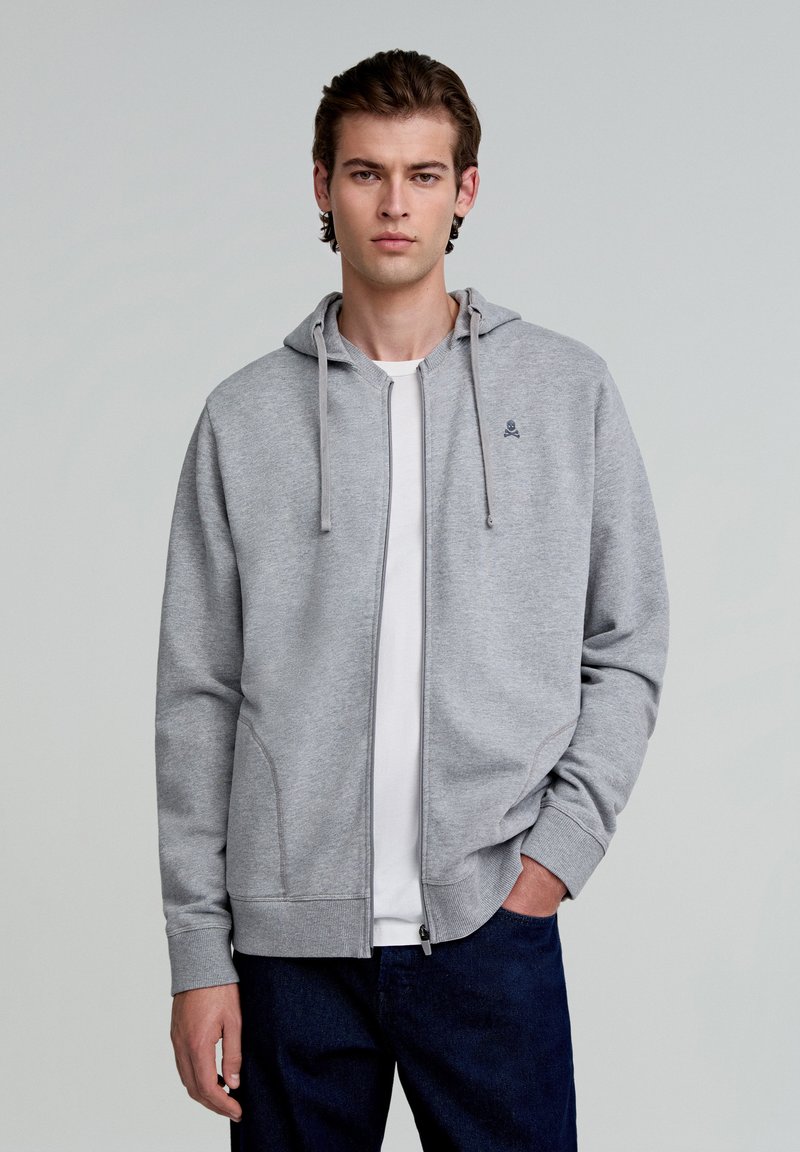Grauer Reißverschluss-Hoodie mit einer Kapuze mit Kordelzug, gerippten Bündchen und Taschen. Wird über einem weißen T-Shirt getragen und mit dunklen Jeans kombiniert. Einfaches Logodesign.