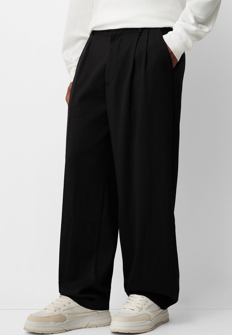 Personne portant un pantalon noir à jambes larges, un haut blanc à manches longues et des baskets écru, debout les mains dans les poches.