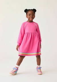 Abito di cotone rosa con maniche lunghe, vita arricciata e ricami arcobaleno. Presenta strisce multicolori all'orlo ed è abbinato a sneakers colorate.