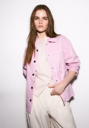 Mladá žena s dlhými hnedými vlasmi, oblečená v svetloružovom prepínacom kabáte cez pruhované tričko a krémové nohavice s širokými nohavicami, stojaca oproti bielej stene.