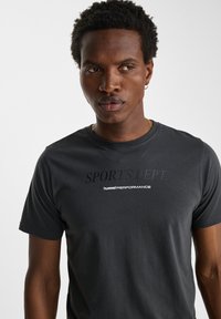 Svart t-shirt med kort ärm i bomull. Har "SPORTS DEPT." och "hummel PERFORMANCE" tryckt i kontrasterande text på bröstet.