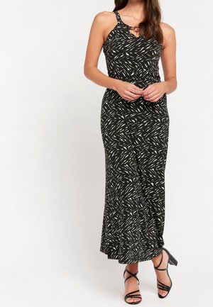 Vrouw draagt een zwarte maxi-jurk met witte en olijfgroene abstracte patronen, dunne bandjes en zwarte haksandalen met riempjes.