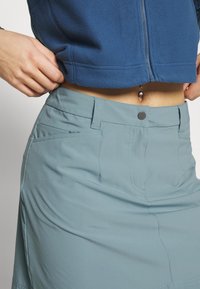 Osoba v světle modré sukni s předními knoflíky a modré zipové bundě, ukazující břicho s piercingem v pupíku.