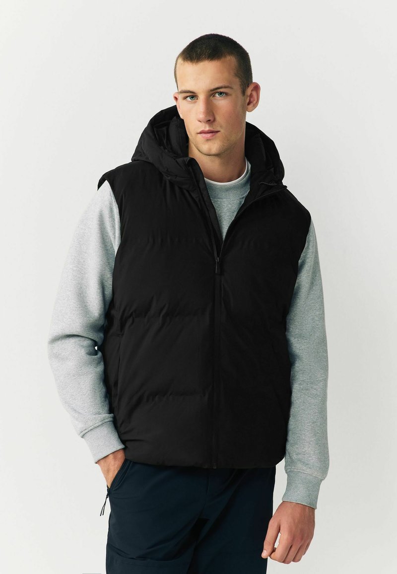 Schwarze Pufferweste mit Kapuze, Reißverschluss und seitlichen Taschen über einem grauen Sweatshirt. Die Weste hat eine gesteppten Textur.
