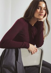 Burgunderfarbene Langarmbluse mit taillierter Passform, getragen mit grauer Outerwear, sitzend auf einem schlichten, metallischen Stuhl. Glatte Textur, lässiger Look.