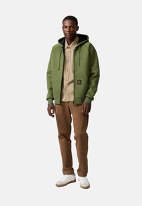 Felpa verde con zip e cappuccio, dotata di tasche frontali e patch con logo, abbinata a una camicia beige con bottoni e pantaloni marroni. Sneakers bianche.