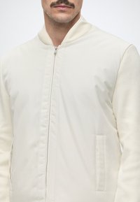 Homme portant une veste bomber beige clair avec une fermeture éclair devant, un col côtelé, des manches texturées et des poches latérales visibles.
