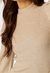 Pull en maille côtelée beige avec un col montant, présentant une texture subtile et des détails de couture sur les épaules et le corps.