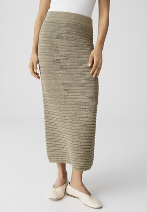 Donna con gonna midi beige a coste in maglia abbinata a ballerine bianche senza lacci, in piedi contro uno sfondo chiaro e semplice.