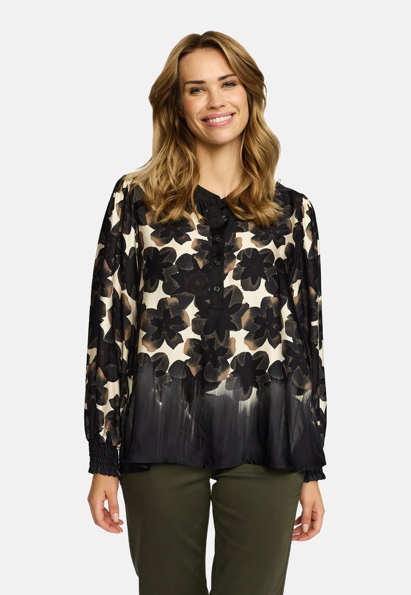 Kvinne smilende, iført en svart og beige blomstret bluse med lange ermer og olivengrønne bukser, stående mot en ensfarget bakgrunn.