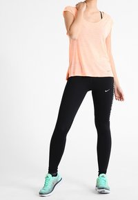 Top de manga corta color naranja claro, leggings negros y zapatillas deportivas multicolor. Presenta logotipos de Nike en la ropa y las zapatillas.