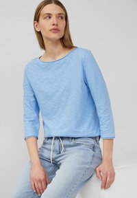 Helles blaues, langärmliges Oberteil mit einem Bootsausschnitt und einem weichen, strukturierten Stoff. Kombiniert mit hellblauen Jeans, die durch einen Kordelzug akzentuiert werden.