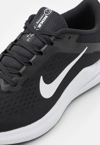 Chaussure de course Nike noire avec logo swoosh blanc, empeigne tricotée, lacets noirs et semelle intermédiaire blanche rembourrée, inclinée pour montrer les détails latéraux et de la languette.