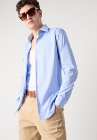 Camisa de botão azul claro feita de um tecido suave, com gola, mangas longas e um corte relaxado, combinada com calças bege e um cinto castanho.