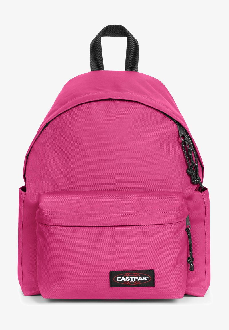 Różowy plecak z materiału o zaokrąglonym wierzchołku, z przednią kieszenią na zamek, czarnymi akcentami oraz łatką z logo na dole. Posiada uchwyt u góry oraz regulowane szelki.
