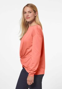 Koraalkleurig shirt met lange mouwen, een losse pasvorm en een geknoopt detail op de taille. Glad textuur en ronde halslijn, gecombineerd met donkere broeken.