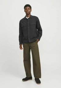 Camisa negra de manga larga con dos bolsillos en el pecho, combinada con pantalones anchos de color verde oliva. Modelo usando zapatos oscuros, de pie frente a un fondo claro.