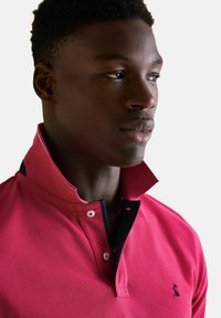 Mladý černoch ve světlém růžovém polo tričku s vystrčeným límečkem, mírně se dívající doprava na pozadí jednoduchého bílého pozadí.