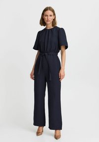Ufficio blu navy con maniche a sbuffo corte, cintura annodata in vita, pantaloni larghi e tessuto testurizzato. Abbinato a tacchi alti color sabbia.