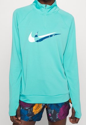 Osoba w turkusowej bluzie z suwakiem do połowy dekoltu, z białym i niebieskim logo w kształcie warstwowego znaku Swoosh oraz w krótkich spodenkach w kwiatowy wzór z żółtymi, niebieskimi i fioletowymi różami.