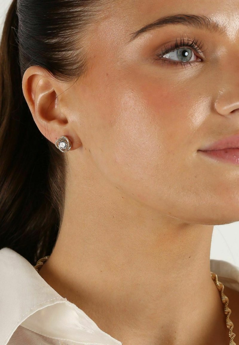 Silverfärgade studs har en rund, klar ädelsten i mitten, omgiven av en texturerad silverring. Modellen bär en ljus blus.