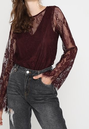 Top en dentelle bordeaux avec de longues manches transparentes ornées de motifs floraux et un décolleté en V, associé à un jean foncé à taille haute.