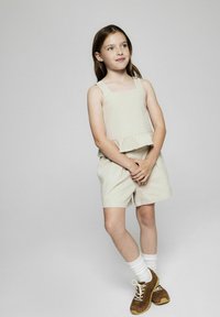Beige, ärmlös topp med volang i nederkanten som matchas med kortbyxor. Lätt tyg med en slät textur. Bruna skor med vita strumpor.