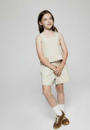Top beige sin mangas con un dobladillo acampanado combinado con pantalones cortos a juego. Tela ligera con una textura suave. Zapatos marrones con calcetines blancos.