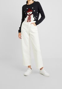 Maglione lavorato a maglia di colore nero con un design di pupazzo di neve, cappello e sciarpa rossi, abbinato a pantaloni bianchi a gamba larga e sneaker bianche.