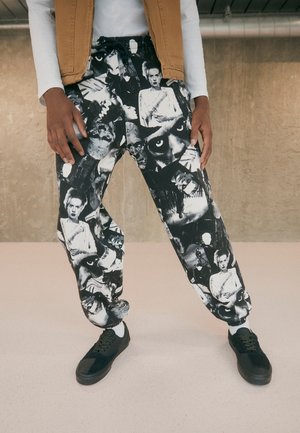 Personne portant un pantalon à imprimé graphique noir et blanc avec des visages et des yeux, associé à des baskets noires, des chaussettes blanches, une chemise blanche et un gilet marron.