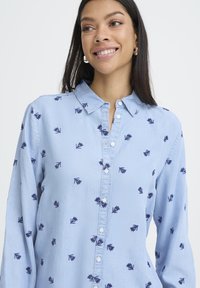 Kvinne med langt mørkt hår som smiler, iført en lyseblå skjorte med knapper og et lite mørkt bladmønster, sølv hoop øredobber, på en hvit bakgrunn.