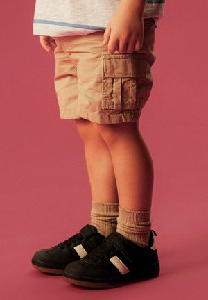 Bambino che indossa pantaloncini beige cargo, calzini marrone chiaro e sneaker nere con strisce bianche, in piedi davanti a uno sfondo rosa.