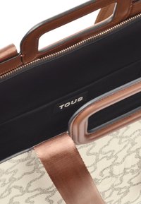 Bolso en tonos beige y marrón con un patrón abstracto, correa de tela marrón, asas en forma de corte rectangular y etiqueta interior negra que dice "TOUS".