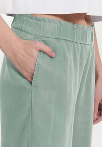 Lichtgroene broek met een elastische tailleband, zijzak en zachte, gestructureerde stof. Een hand rust in de zak.